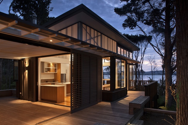 The modest Pahi Bach in Northland was built in 2012. Inserted between trees and wrapping around the path to the harbour edge, the home glows at night for maximum welcome.
