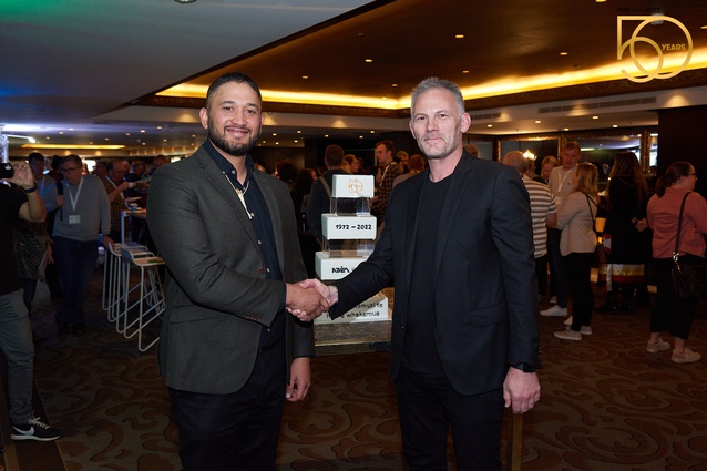 Kīngi Makoare (pictured left) shakes hands with NZILA's President Henry Crothers (right), in front of a four-tier birthday cake marking 50 years of the Institution in Aotearoa.